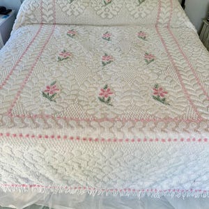 May include: White crochet bedspread with pink floral embroidery and a white lace trim. The bedspread is on a black metal bed frame.