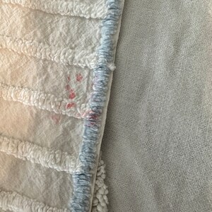 May include: Close-up of a white textured fabric with a blue trim. The fabric has a few small pink stains.
