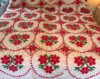 May include: A white bedspread with a repeating pattern of red and green floral hearts. The bedspread has a textured, tufted appearance and is trimmed with a red fringe. The bed frame is black with gold accents.