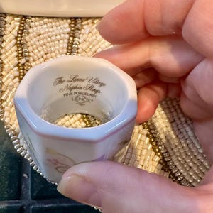 K&ouml;nnte beinhalten: Ein wei&szlig;er Porzellan-Serviettenring mit sechseckiger Form. Der Ring hat einen rosa Rand und goldene Blumenakzente. Der Text "The Lenox Village Napkin Rings FINE PORCELAIN LENOX" ist aufgedruckt.