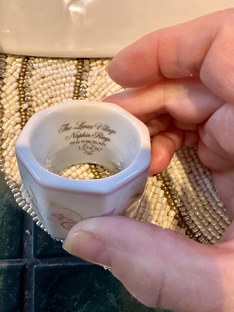 K&ouml;nnte beinhalten: Ein wei&szlig;er Porzellan-Serviettenring mit sechseckiger Form. Der Ring hat einen rosa Rand und goldene Blumenakzente. Der Text "The Lenox Village Napkin Rings FINE PORCELAIN LENOX" ist aufgedruckt.