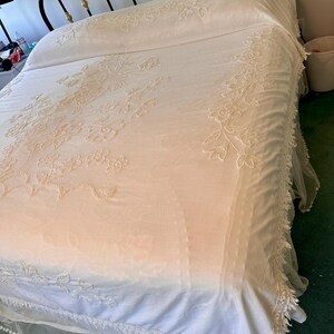 May include: A bed with a black metal frame and gold accents. The bed is covered with a cream-colored bedspread and pillowcases, featuring intricate floral embroidery. A white, lacy trim adorns the edges of the bedspread, adding a delicate touch.