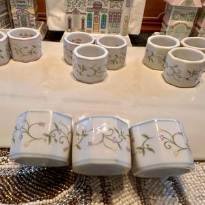 K&ouml;nnte beinhalten: Eine Sammlung wei&szlig;er Keramik-Serviettenringe mit floralem Design. Die Ringe haben einen hellblauen Rand und sind mit gr&uuml;nen Ranken, lila Blumen und gelben Akzenten verziert. Weitere dekorative Keramikteile befinden sich im Hintergrund.