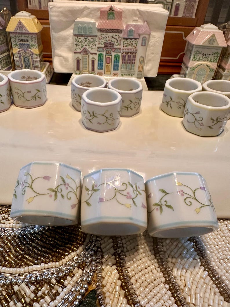 K&ouml;nnte beinhalten: Eine Sammlung wei&szlig;er Keramik-Serviettenringe mit floralem Design. Die Ringe haben einen hellblauen Rand und sind mit gr&uuml;nen Ranken, lila Blumen und gelben Akzenten verziert. Weitere dekorative Keramikteile befinden sich im Hintergrund.