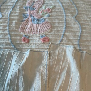 May include: A white crocheted blanket with a blue and pink embroidered design of a girl holding a flower. The blanket has a striped pattern and a blue border.