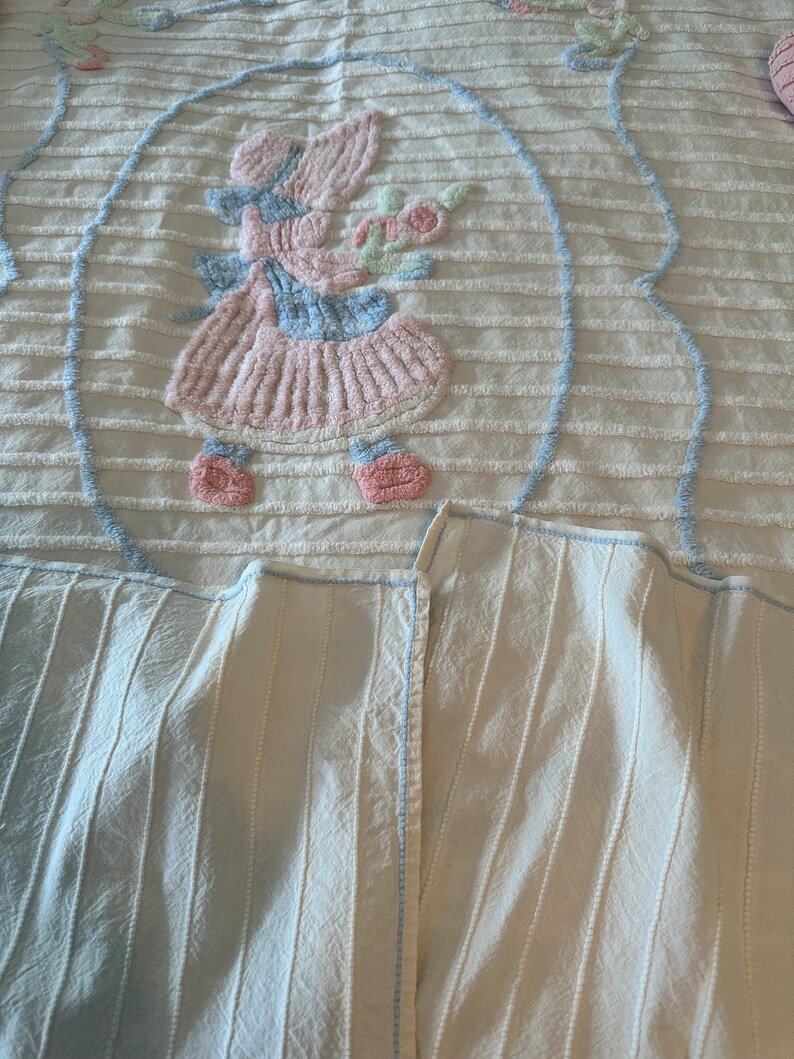 May include: A white crocheted blanket with a blue and pink embroidered design of a girl holding a flower. The blanket has a striped pattern and a blue border.