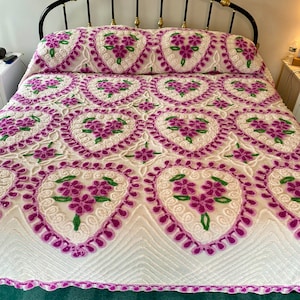 May include: White and purple floral bedspread with a ruffled edge. The bedspread features a heart-shaped design with purple flowers and green leaves.