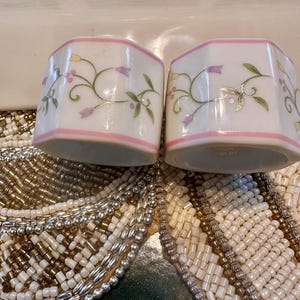 K&ouml;nnte beinhalten: Zwei wei&szlig;e Keramik-Serviettenringe mit rosa Rand und floralem Design. Das florale Design umfasst lila und gelbe Blumen mit gr&uuml;nen Bl&auml;ttern. Die Serviettenringe sind sechseckig.