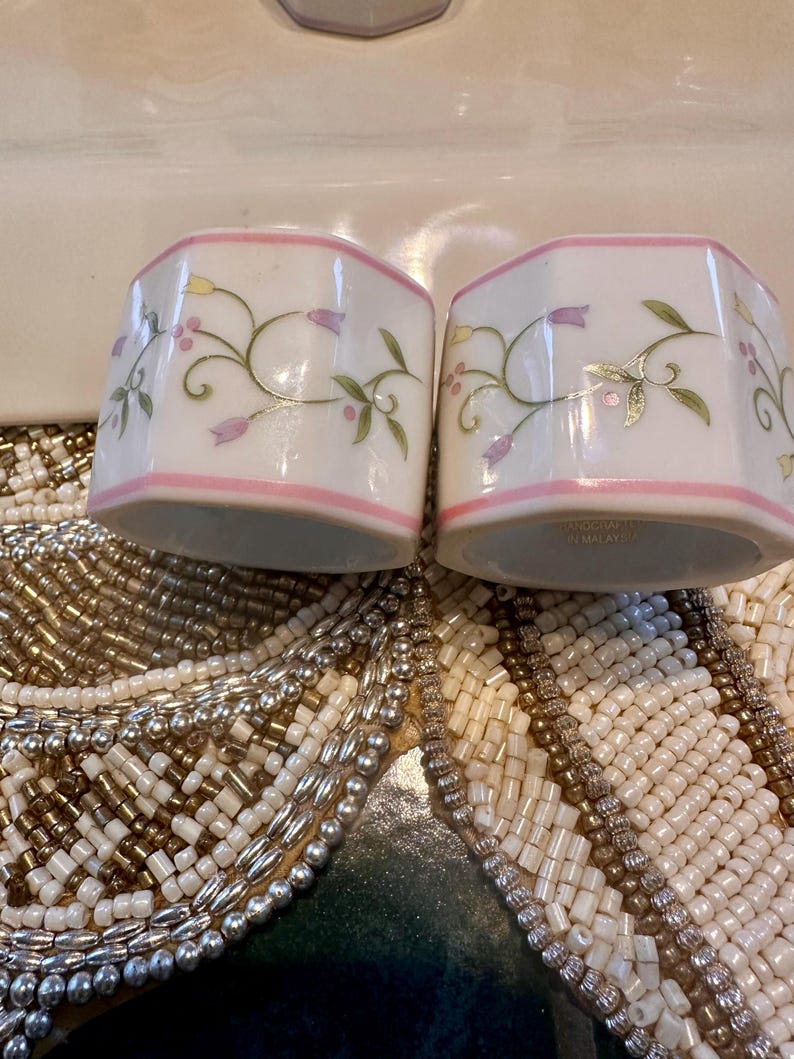 K&ouml;nnte beinhalten: Zwei wei&szlig;e Keramik-Serviettenringe mit rosa Rand und floralem Design. Das florale Design umfasst lila und gelbe Blumen mit gr&uuml;nen Bl&auml;ttern. Die Serviettenringe sind sechseckig.
