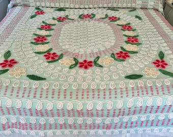 May include: A vintage, light green bedspread with a white and pink floral design. The bedspread has a raised, textured pattern and a white ruffle around the bottom.