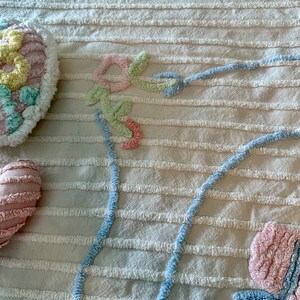 May include: A white textured fabric with a blue, pink, and green floral design. The design is made of fluffy yarn and is stitched onto the fabric.