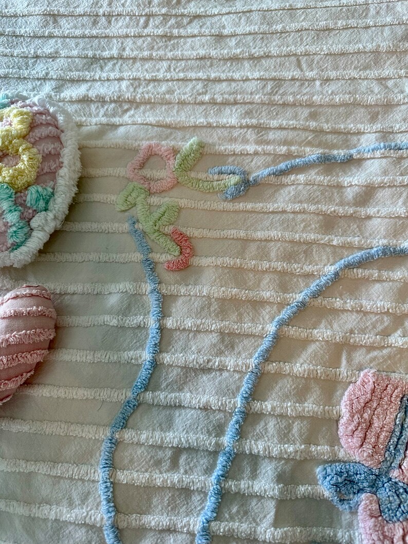 May include: A white textured fabric with a blue, pink, and green floral design. The design is made of fluffy yarn and is stitched onto the fabric.
