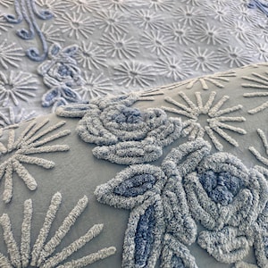 May include: Close-up of a light blue bedspread and pillow with raised floral and starburst patterns. The designs are in varying shades of blue, creating a textured, three-dimensional effect. The bedding appears soft and inviting, suitable for a bedroom.