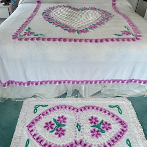 May include: White bedspread and matching rug set with a heart-shaped design. The bedspread features a large heart in the center, surrounded by pink and green floral accents. The rug mirrors the heart design with similar colors and fringe.