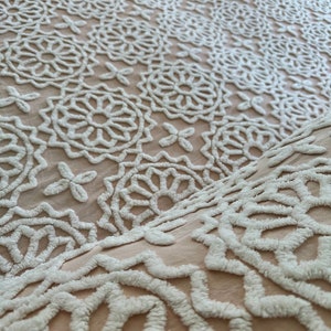 May include: A close-up of a white and beige crocheted throw blanket with a floral pattern.