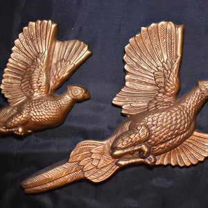 May include: Two gold-colored metal wall sculptures of flying pheasants. The pheasants are in mid-flight with their wings spread out. The sculptures are detailed with textured feathers and have a realistic appearance.