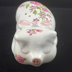 May include: A white ceramic pig figurine with pink and purple floral details. The pig is lying down with its eyes closed and has a pink bow around its neck.