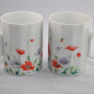 May include: Two white ceramic mugs with a floral design. The mugs have a geometric shape and feature a pattern of red, purple, and green flowers.