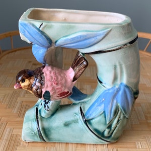 May include: A ceramic planter shaped like a bamboo stalk with a bird perched on a branch. The planter is painted in shades of green, blue, and pink. The bird has brown, pink, and white details.