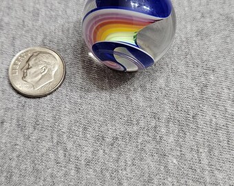 May include: A clear glass marble with a rainbow swirl design. The marble has a blue, white, pink, yellow, and green swirl pattern.