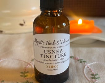 Könnte beinhalten: Eine braune Glasflasche mit Usnea-Tinktur und einer schwarzen Pipette. Das Etikett lautet "Mystic Herb & Thread" und "USNEA TINCTURE". Die Flasche enthält 30 ml Flüssigkeit. Die Flasche steht auf einem floral bestickten Stoff.