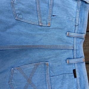 K&ouml;nnte beinhalten: Hellblaue Jeans mit orangefarbenen N&auml;hten. Die Jeans hat zwei Ges&auml;&szlig;taschen, eine mit einem X-f&ouml;rmigen Nahtdesign und die andere mit einem vertikalen Nahtdesign. Das Wrangler-Markenetikett ist sichtbar.