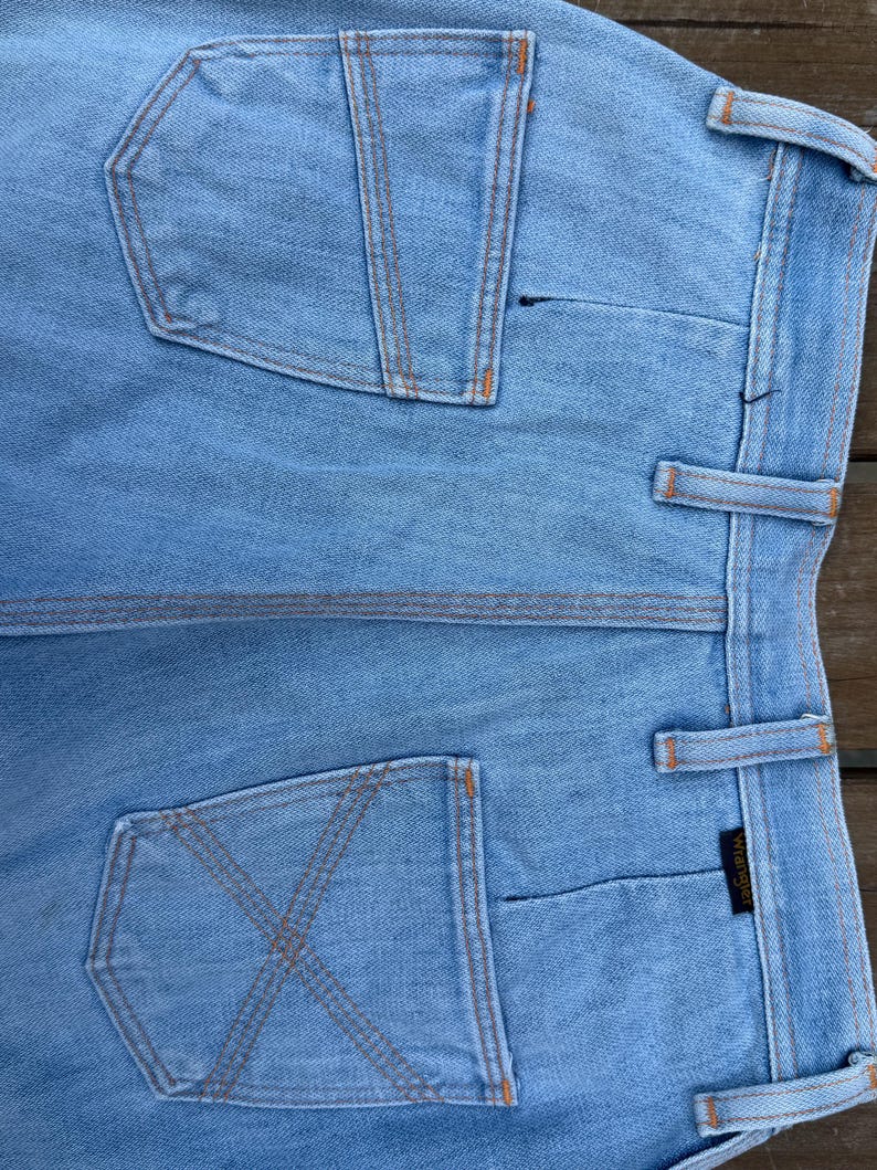 K&ouml;nnte beinhalten: Hellblaue Jeans mit orangefarbenen N&auml;hten. Die Jeans hat zwei Ges&auml;&szlig;taschen, eine mit einem X-f&ouml;rmigen Nahtdesign und die andere mit einem vertikalen Nahtdesign. Das Wrangler-Markenetikett ist sichtbar.