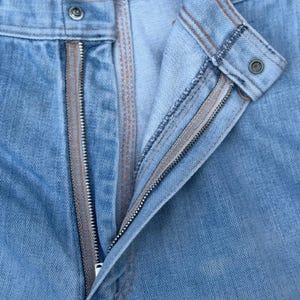 K&ouml;nnte beinhalten: Nahaufnahme einer hellblauen Jeans mit offenem Rei&szlig;verschluss. Die Jeans hat einen Knopfverschluss und braune Nahtdetails. Der Rei&szlig;verschluss ist silberfarben und der Stoff ist leicht verblichen.