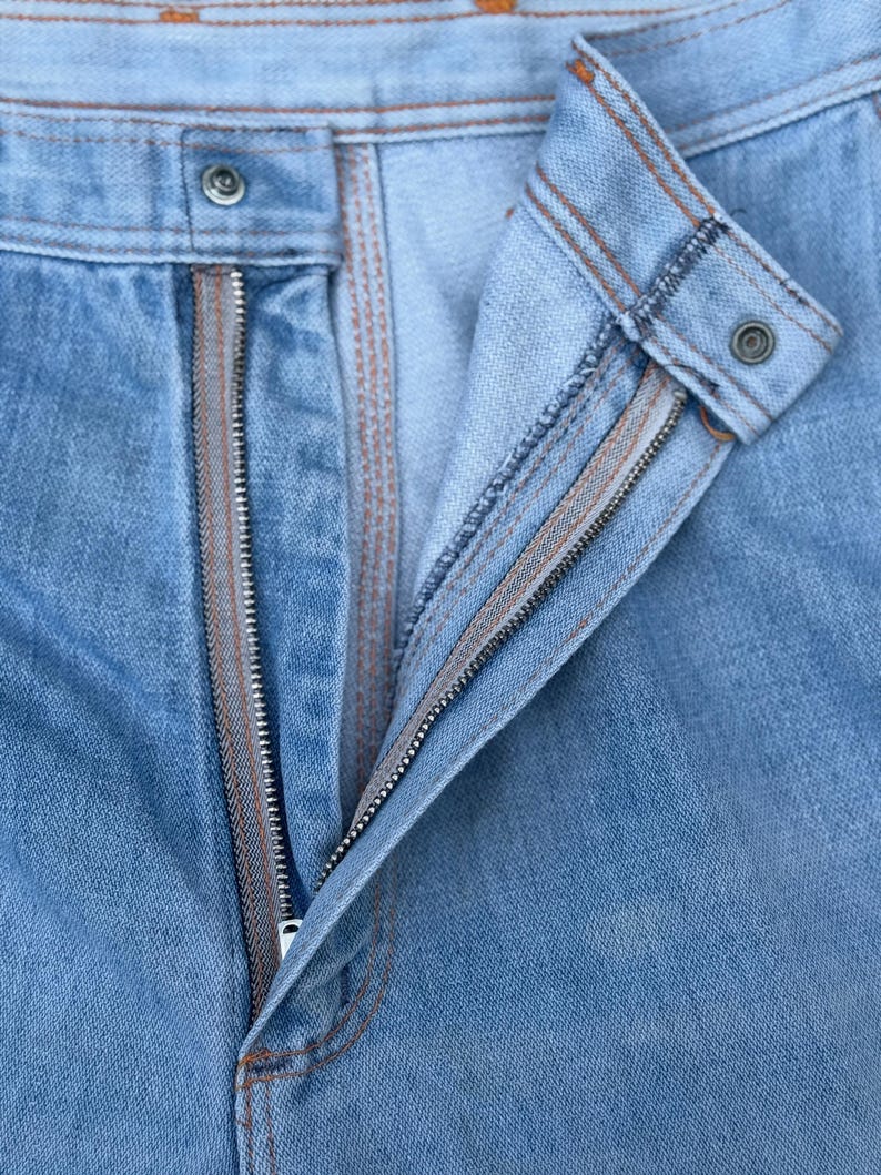 K&ouml;nnte beinhalten: Nahaufnahme einer hellblauen Jeans mit offenem Rei&szlig;verschluss. Die Jeans hat einen Knopfverschluss und braune Nahtdetails. Der Rei&szlig;verschluss ist silberfarben und der Stoff ist leicht verblichen.