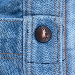 K&ouml;nnte beinhalten: Nahaufnahme von hellblauem Denim-Stoff mit orangefarbenen N&auml;hten und einem bronzefarbenen Knopf. Der Knopf ist mit einem Design gepr&auml;gt. Der Denim hat einen klassischen, abgenutzten Look.