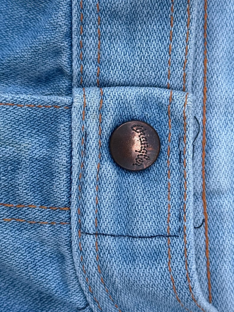 K&ouml;nnte beinhalten: Nahaufnahme von hellblauem Denim-Stoff mit orangefarbenen N&auml;hten und einem bronzefarbenen Knopf. Der Knopf ist mit einem Design gepr&auml;gt. Der Denim hat einen klassischen, abgenutzten Look.
