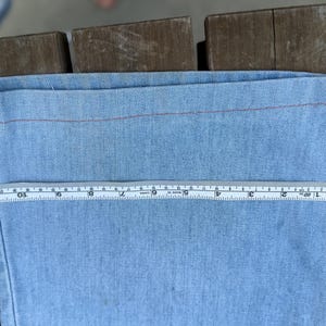 K&ouml;nnte beinhalten: Hellblauer Denimstoff mit roter Naht. Ein gelbes und wei&szlig;es Ma&szlig;band zeigt eine Messung von etwa 30 cm &uuml;ber dem Stoff. Der Stoff liegt auf einer Holzoberfl&auml;che.