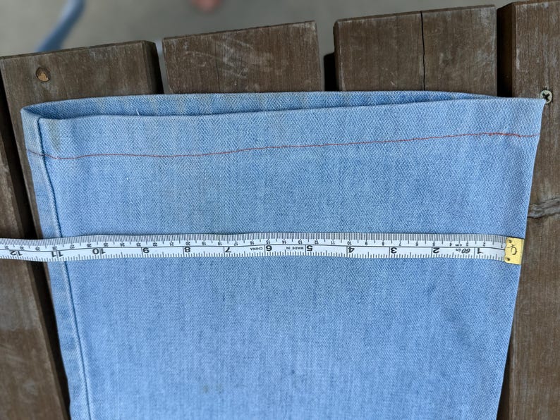K&ouml;nnte beinhalten: Hellblauer Denimstoff mit roter Naht. Ein gelbes und wei&szlig;es Ma&szlig;band zeigt eine Messung von etwa 30 cm &uuml;ber dem Stoff. Der Stoff liegt auf einer Holzoberfl&auml;che.