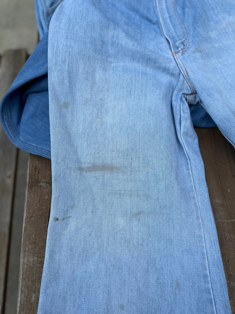 K&ouml;nnte beinhalten: Hellblaue Jeans mit sichtbaren Gebrauchsspuren und Flecken. Die Jeans sind verblichen und weisen Gebrauchsspuren auf. Das Bild zeigt den oberen Teil der Jeans, einschlie&szlig;lich der Taille und eines Teils der Beine. Der Denim scheint ein klassischer, Vintage-Stil zu sein.