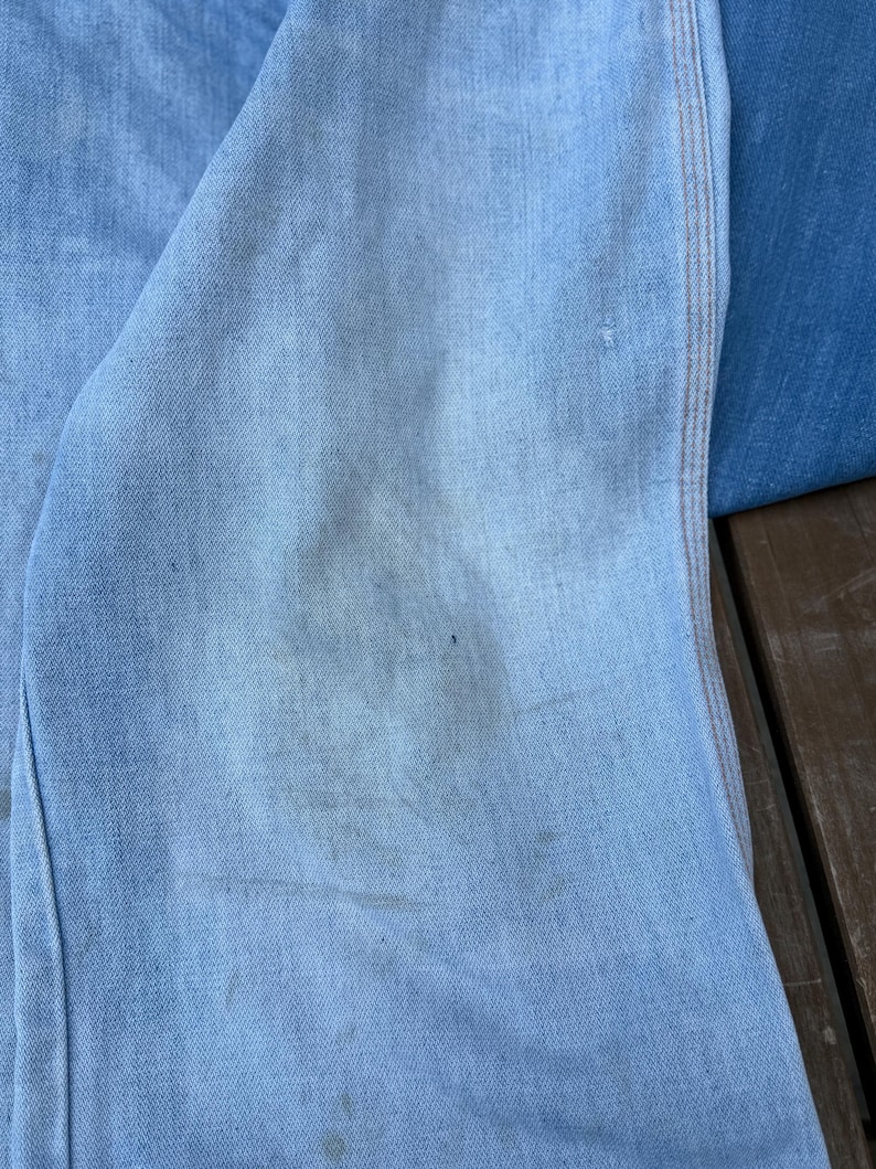 K&ouml;nnte beinhalten: Nahaufnahme von hellblauem Denim-Stoff mit sichtbarer Textur und leichter Verf&auml;rbung. Das Bild zeigt einen Abschnitt aus Denim, m&ouml;glicherweise von Jeans oder einer Jacke, mit einer Naht und Nahtdetails.