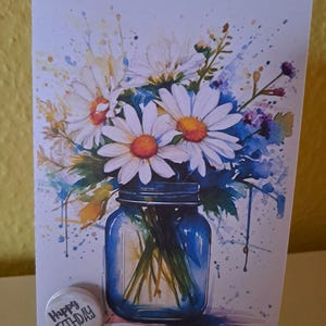 Könnte beinhalten: Ein Aquarellbild eines Straußes weißer Gänseblümchen in einem blauen Einmachglas. Der Hintergrund ist hellblau mit weißen Farbspritzern. Die Karte hat einen kleinen runden Aufkleber mit der Aufschrift "Happy Birthday".