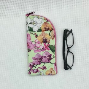 May include: A light green glasses case with a pink zip and trim, decorated with a floral orchid pattern in pink, white, and orange. Black-framed glasses are placed next to the case.