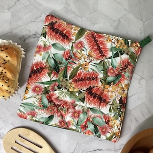 May include: A square quilted potholder featuring a botanical print of red bottlebrush flowers, green leaves, and small yellow buds on a white background, with a small green fabric loop attached to one corner for hanging in the kitchen.