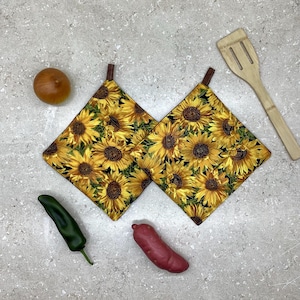 May include: Two square pot holders with a sunflower design, showing yellow and brown flowers on a black backdrop. Each has a brown loop. A wooden spatula, a tomato, a pepper, and a sweet potato are also visible.