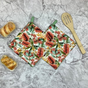 May include: Two square pot holders with a floral design of red, yellow, and green botanical elements on a white backdrop. Each has a green hanging loop. A wooden spoon and two rolls are also in the image.