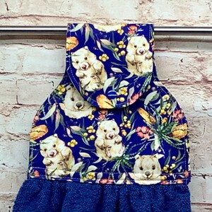 May include: A blue kitchen towel with a hanging loop features a whimsical pattern of wombats and wattle flowers on a dark blue background. The towel has a blue terry cloth fringe.