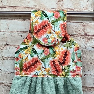 May include: A hanging kitchen towel with a floral pattern featuring red, yellow, and green flowers on a white background. The bottom portion of the towel is a solid light green colour. The towel is hanging on a metal bar against a brick wall.