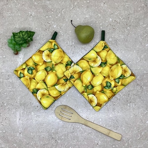 May include: Two yellow lemon-patterned pot holders with green trim and a green hanging loop. The pot holders are on a grey countertop with a wooden spoon.