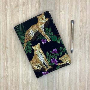 May include: A black notebook cover with a leopard print design. The cover features three leopards, green leaves, and purple flowers. The notebook cover has a button closure.