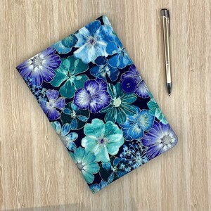 May include: A book cover with a floral pattern in blue and teal hues, secured with a button. A silver pen lies next to the cover on a light wood surface. The cover's design features repeating flowers.