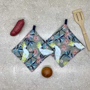 May include: Two grey and white pot holders with a floral pattern and white cockatoos. The pot holders have a dark blue trim.