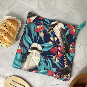 May include: A square quilted potholder featuring a dark blue background with a repeating pattern of kookaburra birds, green eucalyptus leaves, and red flowering gum blossoms. The fabric has a stitched diamond pattern and a small fabric loop corner.