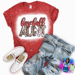 May include: Red t-shirt with a white bleached design that says "baseball aunt" with a leopard print and baseballs. The shirt is on a white background with a pair of blue jeans and white sneakers.