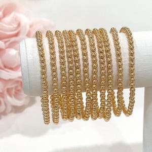 May include: A collection of gold beaded bracelets displayed on a white cylindrical stand. The bracelets are made of small, evenly spaced, round beads, creating a textured surface. The background features soft pink floral elements.