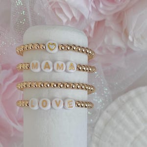 May include: Four gold beaded bracelets with white letter beads spelling out "LOVE" and "MAMA" and a heart charm.